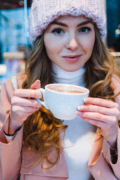 Cup of hot cocoa. Portrait of beautiful young woman in pink coat looking at camera and drinking coffee in cafe - Fotoğraf, Görsel