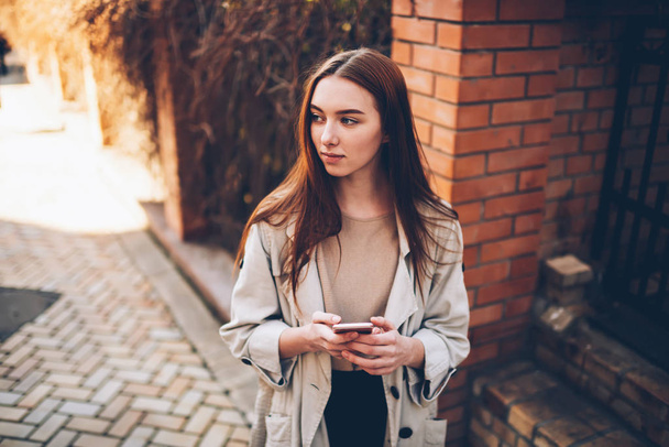 Attractive young female dressed in stylish gray overcoat enjoying  time at urban setting while looking away. charming teenager in good mood relaxing with modern gadget and good mood - Photo, Image