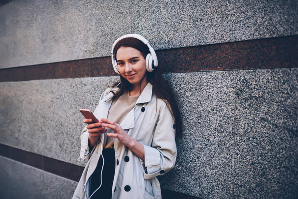Portrait of attractive hipster girl enjoying free time outdoors listening to favorite songs from playlist using application on smartphone and modern headphones standing on promotional background - Foto, immagini