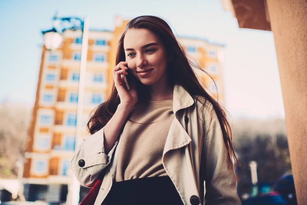 Charming female traveler dressed in trendy casual wear talking on phone using connection in roaming while walking on urban setting background, hipster girl strolling on spring day calling to friend - 写真・画像