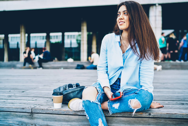 Cheerful hipster girl enjoying favourite songs in earphones connected to smartphone device.Positive student dressed stylish casual outfit listening music in playlist on telephone sitting outdoors - 写真・画像