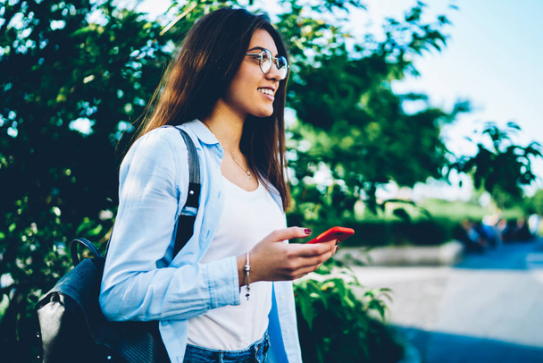 Side view of positive brunette student with backpack enjoyig good weather holding modern telephone in hands.Cheerful hipster girl in optical eyewear checking email on mobile while looking forward - Zdjęcie, obraz