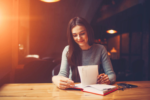 Smiling young woman with brunette hair turning pages and searching useful information in own notepad to preparing for training seminar sitting in coworking with filter effect.Copy space area for text - Zdjęcie, obraz