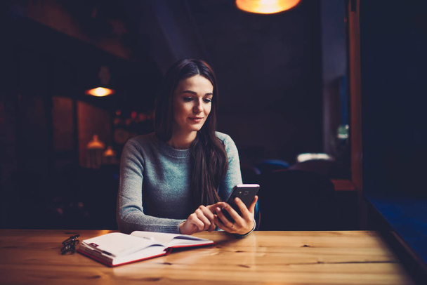 Attractive student sitting at wooden table with notepad and installing new application on modern telephone device connected to 4G internet.Cheerful young woman reading posts in blog on smartphone - Foto, Bild
