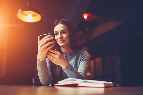 Attractive student watching video in social networks on digital telephone sitting in university interior with training book.Hipster girl reading news on website on modern smartphone in coworking space - Zdjęcie, obraz