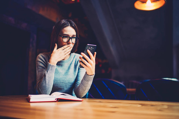 Shocked young woman in optical spectacles reading bad news in social networks on digital smartphone device connected to free 4G internet sitting at book in university interior near copy space area - Фото, изображение