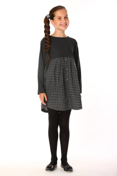 Pretty slender young schoolgirl stands in full growth in uniform. Beautiful attractive little girl posing in business clothes. Cute child is smiling and happy. - Valokuva, kuva