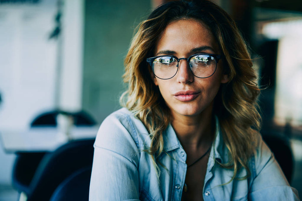 Half length portrait of young attractive woman in trendy eyeglasses enjoying leisure time in coffee shop.Beautiful hipster girl dressed in casual outfit looking at camera while sitting indoors - 写真・画像