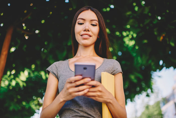 Positive brunette woman reading news from network on mobile spending time in park on summer day,smiling hipster girl checking application notifications on smartphone satisfied with wireless connection - 写真・画像