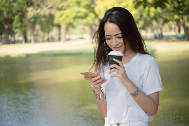 Молодая красивая женщина держит одноразовый кофе чашку в то время как текстовые сообщения через смартфон на природе
. - Фото, изображение