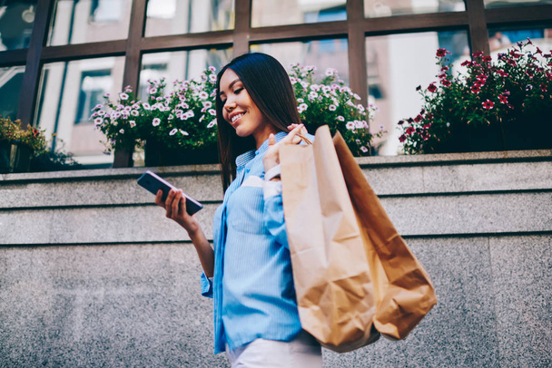 Attractive smiling asian female blogger using personal website online banking via cellphone device walking at urban setting. Young cheerful woman reading messages standing with shopping bags outdoors - Valokuva, kuva