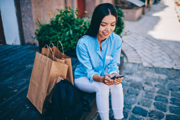 Good looking positive businesswoman sending funny messages to friends via application on smartphone connected to 4G internet.Pretty smiling hipster girl enjoying resting at urban setting with device - Foto, imagen