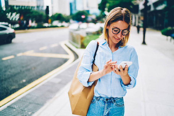 Young woman in eyewear and trendy casual outfit walking on street texting message on smartphone,blonde female  traveler searching location using mobile application for navigating in downtown - Фото, зображення