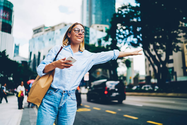 Smiling female traveler in trendy casual wear raising hand calling taxin standing on road in city on urban background, young woman waiting for cab for transporting in megalopolis waving on avenue - Photo, Image