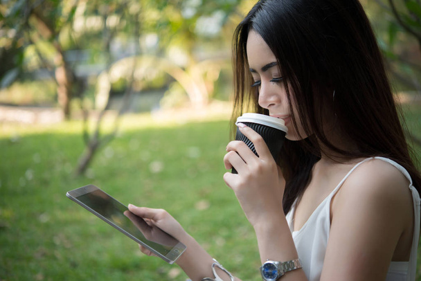 Young beautiful woman holding disposable coffee cup while text messaging through smartphone at nature outdoors. - Photo, Image
