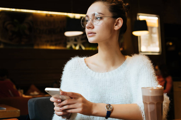 hermosa joven freelancer con gafas sentado en un café, sosteniendo un teléfono inteligente, sobre la mesa es un delicioso cappuccino
 - Foto, Imagen