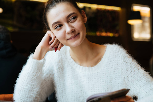schattig jong meisje student in een witte jas zit in een café ambassadeur studeren, glimlachend - Foto, afbeelding