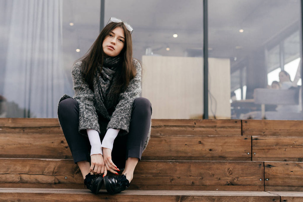tyylikäs tumma tukka nuori tyttö trendikäs harmaa takki istuu portailla, poseeraa
 - Valokuva, kuva