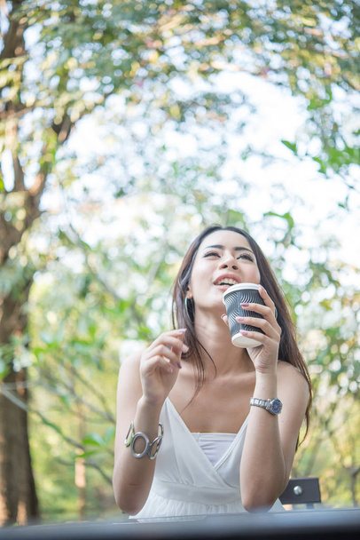Mujer joven sosteniendo taza de café desechable mientras está sentado disfrutando en la naturaleza al aire libre
. - Foto, imagen