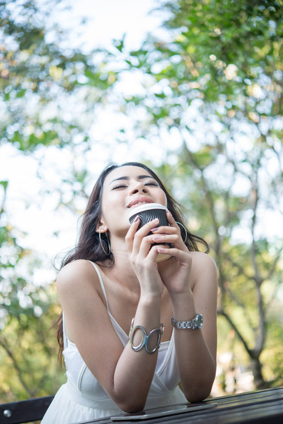Молода жінка тримає одноразову чашку кави, сидячи, насолоджуючись на природі на відкритому повітрі
. - Фото, зображення