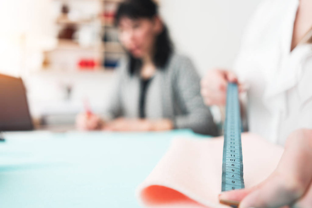 Young dressmaker woman measures the fabric with a ruler. Two seamstress working with cloth in design bureau. Small business. Blurred background - Fotografie, Obrázek