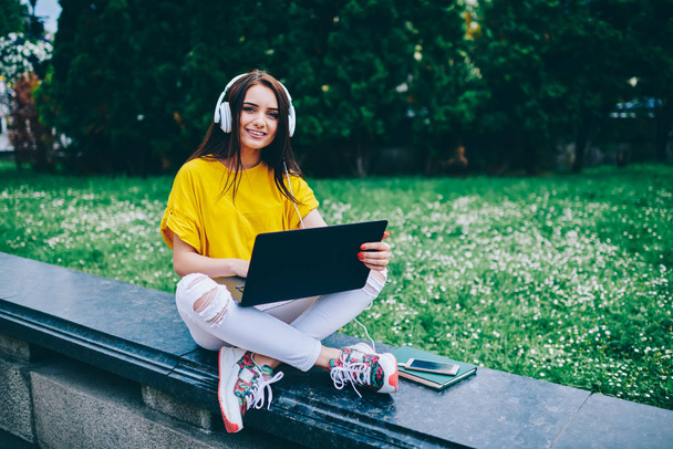 Portrait of cheerful brunette hipster girl in modern headphones connected to modern telephone listening favourite music sitting outdoors with digital laptop computer connected to free wifi internet - Foto, afbeelding