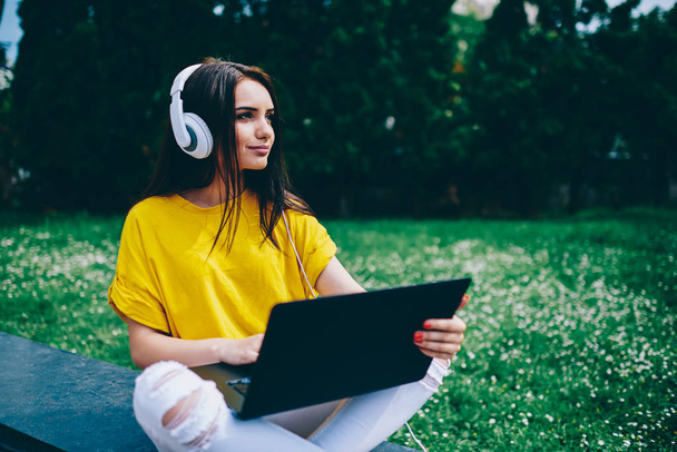 Pondering young woman enjoying favourite music in modern headphones sitting in city centre with digital netbook on green promotional background with publicity space area for advertising area - Фото, зображення
