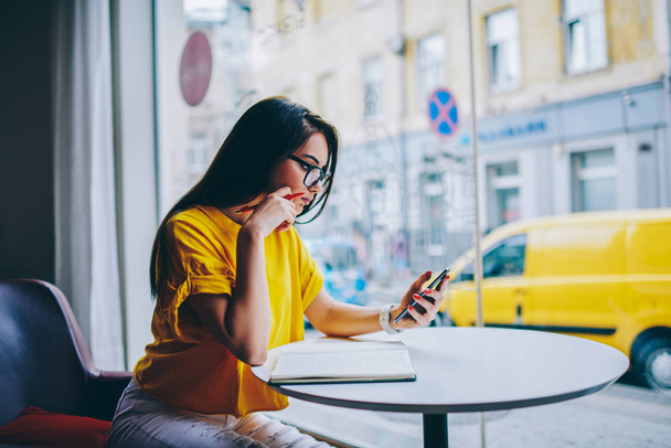Side view of brunette student in eyeglasses reading task of homework on digital telephone to preparing for seminar sitting at table with textbook in coffee shop interior near publicity area - Фото, зображення