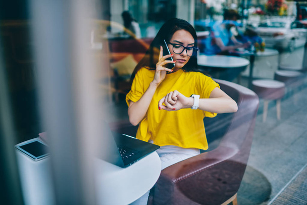 Busy businesswoman in eyewear looking at smartwatch while talking on cellular and agree on meeting sitting at laptop computer and working remotely.Manager managing time during phone conversation - Фото, изображение