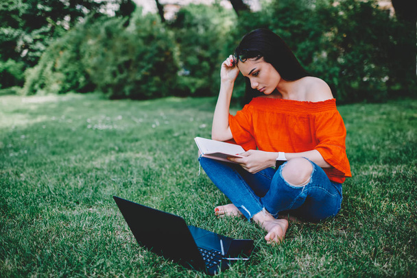 Attractive pondering talented student sitting on grass in university campus and preparing for upcoming examination.Young cute hipster girl planning working schedule using notepad at urban setting - Photo, Image