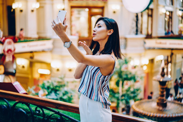 Attractive young woman making air kiss for followers in personal blog shooting video on smartphone, beautiful asian girl posing taking selfie using application on mobile standing in shopping center - Fotó, kép