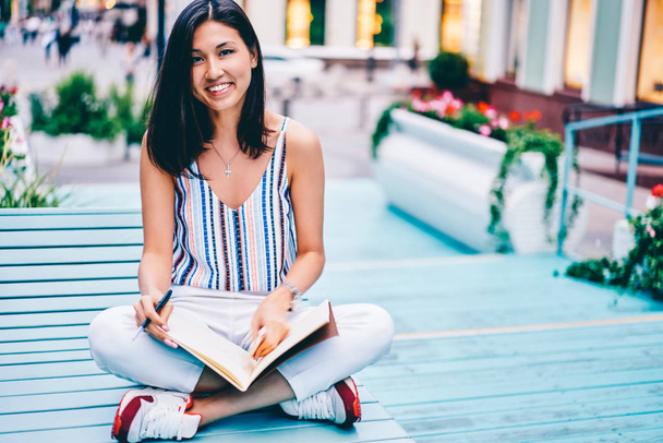 Portrait of cheerful young woman sitting with notebook for writing journey plans recreating outdoors, smiling female student looking at camera creating idea for graphic drawings in sketchbook - 写真・画像
