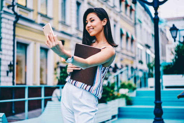 Smiling hipster girl in trendy apparel posing for selfie on smartphone camera, cheerful asian woman making photo on application on mobile for share online while strolling on city street with folder - Foto, Imagem