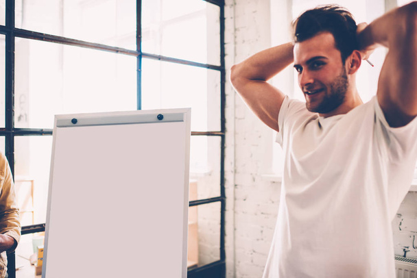 Handsome male student standing near blank board preparing for explaining his ideas for startup, happy hipster guy thinking over making speech and using flip chart for presentation creative solutions - Фото, зображення