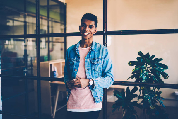 ソーシャル ネットワークで仕事休憩のブログに満足して陽気なアフロ アメリカンの若い男の肖像のオフィスで時間を費やしてのスマート フォンでメールをチェック カメラを見て成功した流行に敏感な男 - 写真・画像