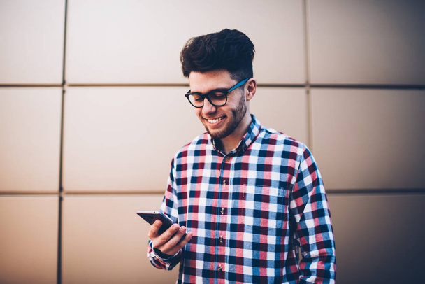Cheerful hipster guy in plaid shirt  happy receiving message from friend on mobile phone, young male student in eyewear checking email on cellular excited with getting discount for sending SMS - Фото, зображення