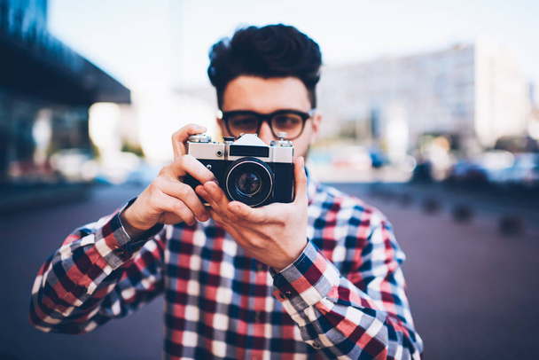 Concentrated hipster guy focusing lens on old vintage camera taking pictures on urban settings, skilled young male amateur in eyeglasses and plaid shirt making photos spending leisure on hobby - Photo, image