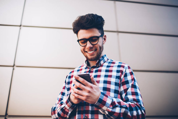 Cheerful male student in eyeglasses blogging in social networks via mobile chatting with followers, smiling hipster guy in plaid shirt receiving photos from friend on email while standing outdoors - 写真・画像