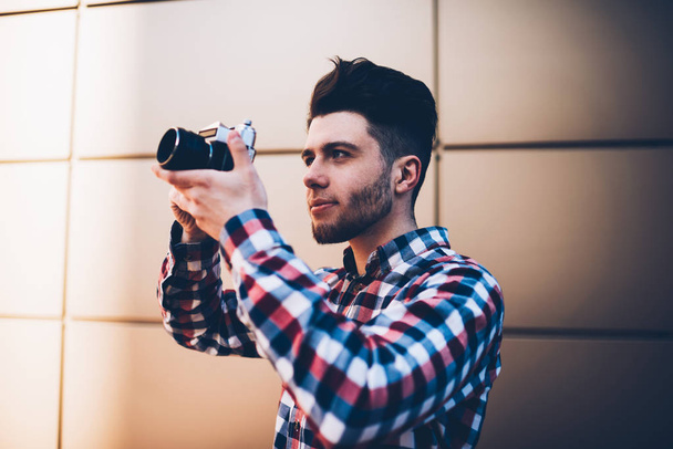 Professional young male photographer taking picture on old camera focusing lens concentrated on work, hipster guy fond of photography using vintage equipment making images standing outdoors - Photo, image