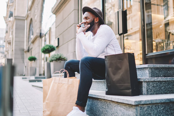 Happy african american male buyer in trendy wear talking on phone with friend about successful shopping, cheerful dark skinned guy having mobile conversation with operator sitting on stairs with bags - Φωτογραφία, εικόνα