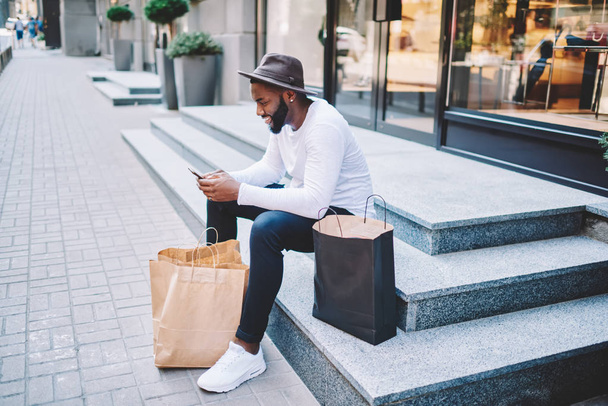 Smiling afro american male customer in trendy hat sitting on stairs of store waiting for delivery service sending email on mobile, dark-skinned guy satisfied with online shopping resting with bags - Foto, immagini