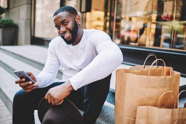 Portrait of african american guy wait for feedback from web store service sitting on stairs wait for purchase,smiling dark-skinned man making payment online via mobile satisfied with online shopping - Foto, immagini