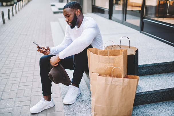 Pensive african american guy using application on phone resting on stairs of store near bags with copy space for brand name or label, dark-skinned man read message on mobile after receiving purchases - Photo, image