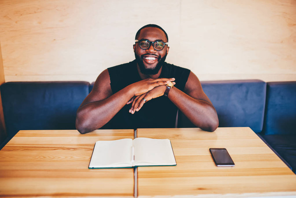 Portrait of cheerful dark skinned businessman in casual wear looking at camera excited with successful startup,afro american guy in eyewear sitting in cafe at wooden table and notepad with blank pages - Foto, Imagen