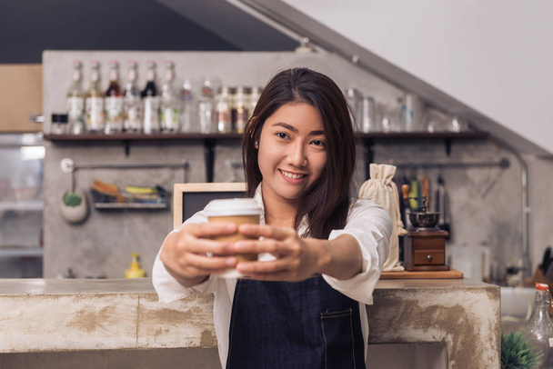 Close up of a young Asian female barista hold a cup of coffee serving to her customer with smile surrounded with bar counter background. Young female barista and her small shop. Food and drink concept - Foto, Imagen