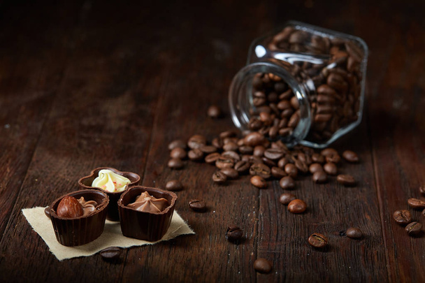 Side view of overturned glass jar with coffee beans and chocolate candies on wooden background, selective focus - Zdjęcie, obraz
