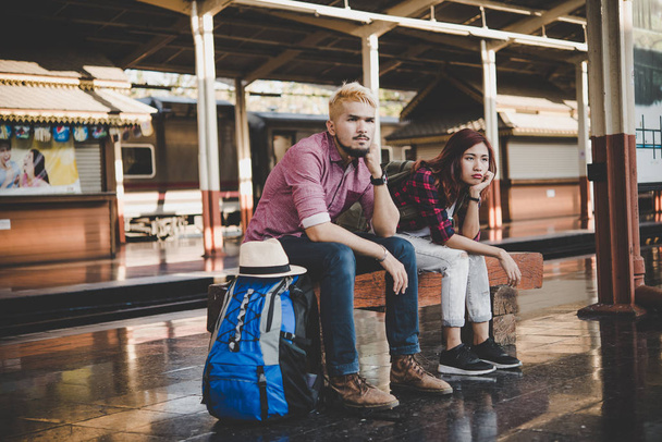 Young hipster couple sitting on wooden bench at train station. Couple sitting waiting for the train at platform. - 写真・画像