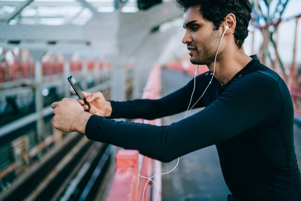 Side view of sportsman dressed in active wear for workout listening music in earphones connected to smartphone holding in hands.Fit man choosing audio app for installing on phone for sport training - Foto, Bild