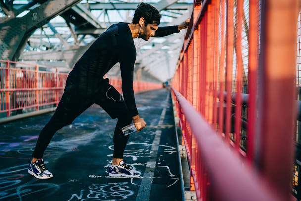 Sportsman with sport goal doing exercises while listening music in earphones for better vitality in urban setting.Fit man dressed in black tracksuit warming up before morning workout on bridge - Foto, Bild