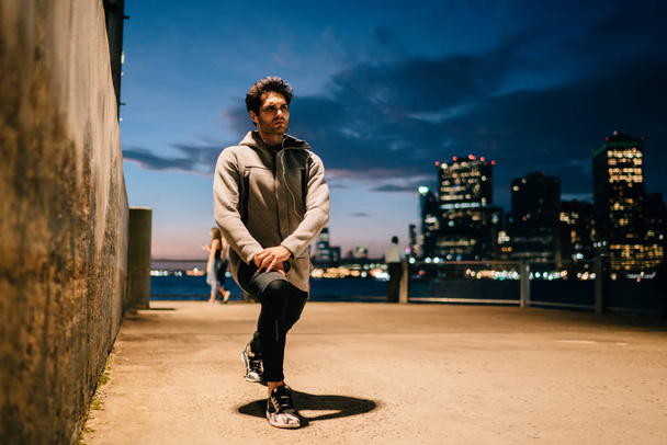 Serious fit man dressed in tracksuit warming up before evening training in urban setting on metropolis promotional background.Young athlete in earphones doing sport exercises outdoors - Foto, imagen